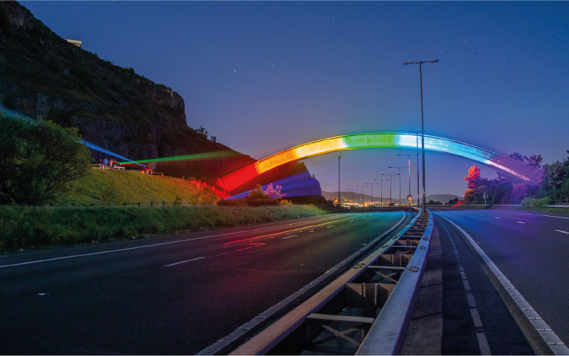 Photograph of a bridge at night lit up with neon rainbow coloured lights