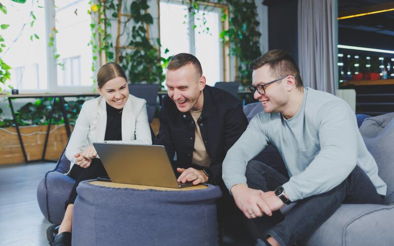 two men and a woman sat in a café on a sofa around a laptop smiling