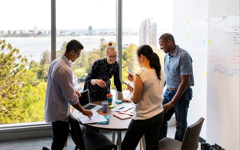 four people standing round a table with a laptop and paper notes, talking about work