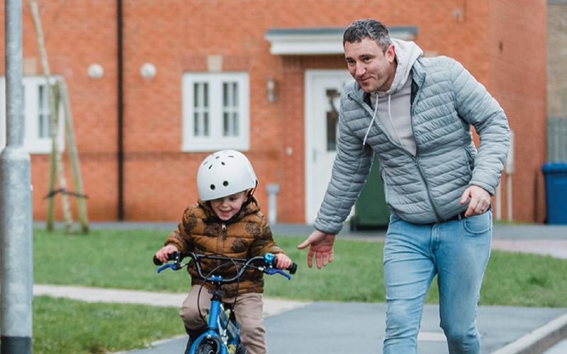 Picture of a father running with his young boy who is riding a bike with houses in the background.