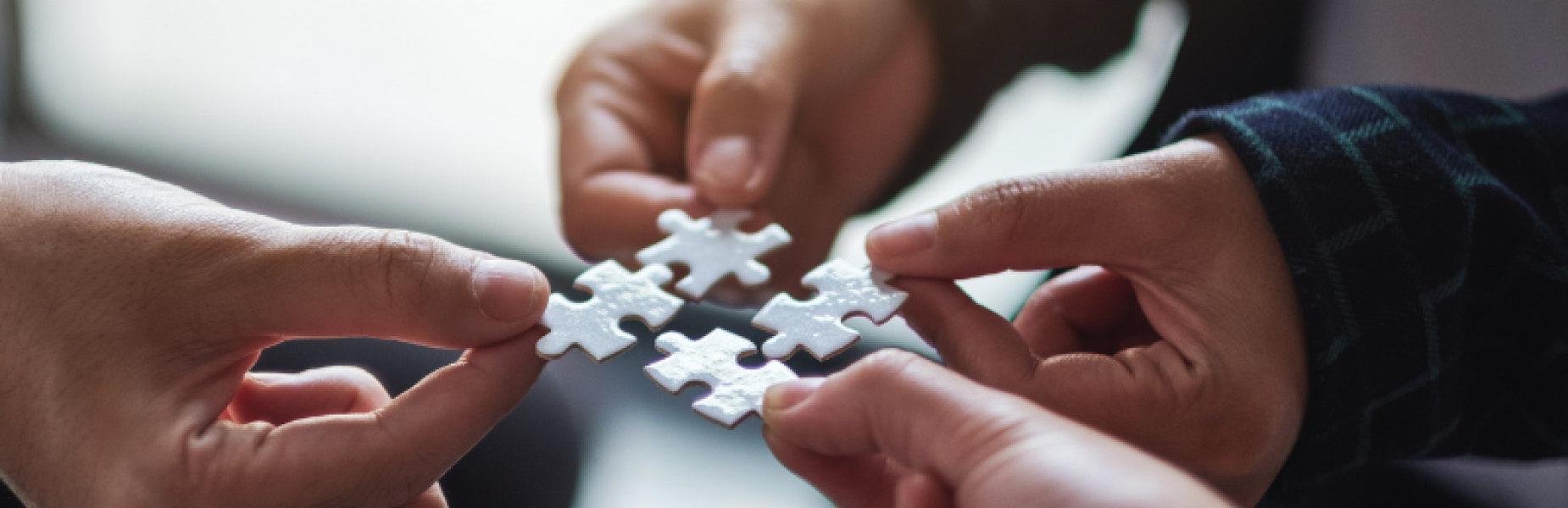 Close‑up of multiple hands holding white jigsaw puzzle pieces and bringing them together, representing teamwork and problem‑solving.