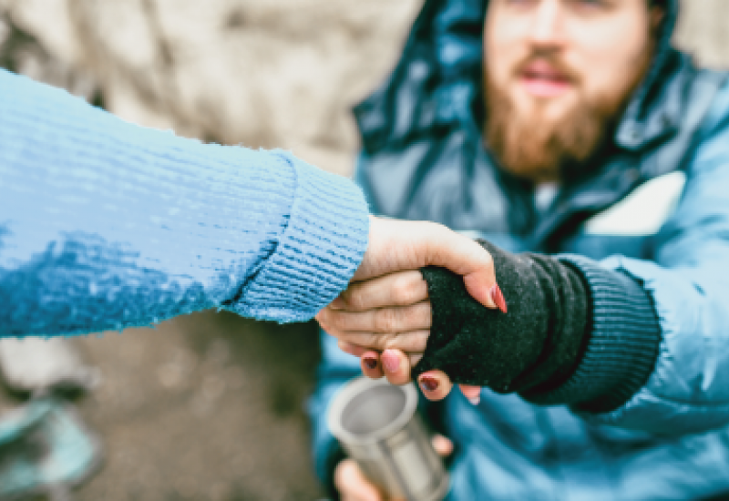 Photograph of an outstretched arm grasping the arm of a seated, bearded homeless man