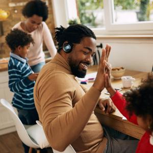 Family sat and stood round the table in the kitchen. The dad is high fiving his child whilst also working. The mum is being shown something by her son. 