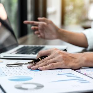 A person reviews charts and reports on a desk while working on a laptop. Papers displaying graphs, charts and performance data are spread across the workspace, with a pen resting on a report.